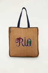 Bolso tote de mano y hombro, color marrón con textura tejida de efecto natural, asas dobles azules y bordado frontal con la palabra 'Rio' decorada con una palmera y un tucán.