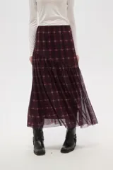 Falda larga de corte evasé con estampado de cuadros escoceses en tonos bordó y negro. Presenta un diseño de doble capa con una base opaca y una sobrecapa de tul semitransparente, con un corte asimétrico en la parte inferior.
