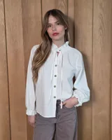 Camisa de color blanco con cuello alto fruncido y lazo para anudar. Presenta cierre frontal con botones de contraste, mangas largas con puños elásticos y terminaciones con volados.