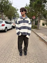 Buzo tipo polo de corte oversize con diseño de rayas horizontales anchas en color gris oscuro y crema. Presenta cuello camisero, cierre frontal parcial con botones ocultos y detalles de costuras expuestas y deshilachadas. Incluye el logo de la marca bordado en el centro del pecho.