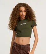 Blusa baby look verde militar de algodón con elastano, cuello redondo, mangas cortas y estampado frontal con la frase "No Time For Love" en color blanco.