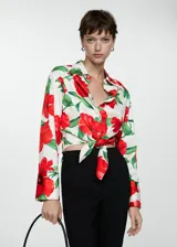 Camisa de corte crop con cuello camisero y cierre frontal de botones, destacada por su diseño estampado de flores rojas sobre fondo blanco. Presenta mangas largas con puños abotonados y un detalle de lazo anudado en la parte inferior delantera.