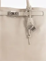 Bolso tipo tote color gris, confeccionado en cuero vacuno, con asas largas para hombro, detalles metálicos personalizados, cierre central y llavero con candado y llave colgante.
