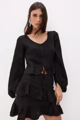 Top negro de lino italiano con escote en V, jareta ajustable en el centro delantero y volado en el ruedo. Mangas largas estilo globo con puños ajustados.