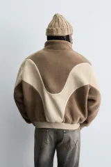Cazadora de corte relajado confeccionada en tejido de borreguillo con diseño de paneles en tonos marrón y beige. Presenta cuello subido, cierre frontal de cremallera, bolsillos laterales y acabados en rib en puños y cintura.