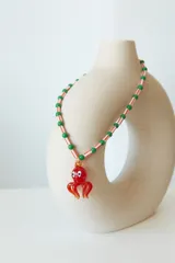 Collar corto de cuentas cilíndricas blancas con rayas naranjas y cuentas esféricas verdes, con un dije colgante de vidrio en forma de pulpo rojo.