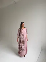 Vestido largo color rosa, con mangas cortas y diseño holgado, presenta un estampado floral en tonos marrones y verdes en un lateral, además de un bolsillo aplicado en la parte frontal y costuras visibles de estilo rústico.