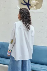 Camisa blanca de corte oversize con bordados de motivos playeros y mangas largas con puños decorados con ondas de colores.