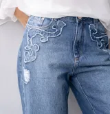 Jean ancho azul con detalles de apliques de tela desflecada en los bolsillos y roturas sutiles en la pierna.