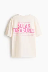 Remera oversized en punto de algodón color beige, de cuello redondo y manga corta, con estampa frontal pequeña en tono rosado con la inscripción "Solar Treasures".