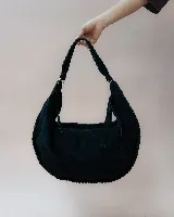 Bolso tipo hobo de gran tamaño, confeccionado en gamuza color marrón oscuro. Presenta un diseño artesanal con costuras visibles y detalles de tientos en el cuerpo, asa integrada con pespuntes a mano y cierre superior.