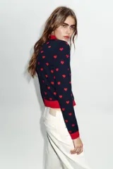 Chaqueta de punto jacquard en color azul marino con estampado de corazones rojos. Presenta cuello de solapa, manga larga y cierre frontal con botones rojos en forma de corazón. Los puños, el cuello y el bajo son de color rojo liso.