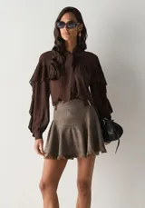 Camisa de color marrón chocolate, confeccionada en tela fluida con diseño de volados en la parte superior y mangas largas abullonadas. Presenta un cuello alto con lazo frontal integrado para anudar.