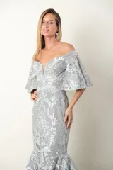 Vestido largo de fiesta color gris perla, corte sirena, con escote off-shoulder y mangas cortas con volados. Confeccionado en tul bordado con hilo al tono y leve brillo.