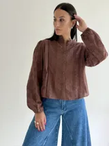 Blusa de brodery color blanco, con cuello redondo, mangas largas abullonadas con puños elásticos y cierre frontal con botones forrados. Presenta detalles de puntillas verticales en el frente.