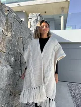 Ruana tejida en lana merino, color beige y crudo, con diseño de trenzas verticales en el frente y terminación con flecos en el ruedo.