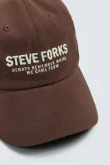 Gorra de seis paneles confeccionada en tejido de algodón, color marrón, con bordado frontal en color crema que incluye texto. Cuenta con ajuste en la parte posterior.