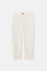 Pantalón tipo jogger de tiro medio, color blanco roto o crema, confeccionado con una mezcla de lino. Presenta bolsillos delanteros y bolsillos de plastrón en la espalda. El bajo tiene pliegues y el cierre es frontal con cremallera y botón.