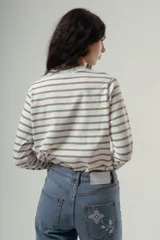 Camiseta de manga larga, cuello redondo, con rayas horizontales marrones sobre fondo blanco. Presenta un pequeño logo bordado en rojo en el pecho.