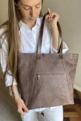 Bolso grande tipo tote color beige con textura tejida y costuras decorativas en los laterales. Tiene doble asa de hombro y logo de la marca en el frente. Interior forrado con bolsillo con cierre y portacelular.