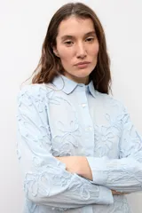 Camisa celeste con apliques florales bordados, cuello camisero, manga larga y cierre frontal de botones.