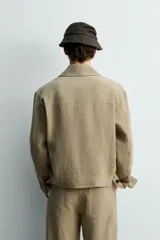 Sobrecamisa cropped fit de lino color beige, con cuello solapa, mangas largas con puño abotonado, bolsillos frontales de vivo y cierre frontal con botones oscuros.