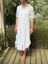 Vestido camisero largo de lino blanco, con cuello camisero, manga corta enrollada y cierre frontal de botones de nácar. Presenta aberturas laterales en el bajo.