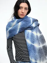 Bufanda de punto grueso tejida en lana, con diseño teñido mediante técnica shibori en tonos azules y blancos, y terminación con flecos en los extremos.