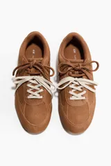 Calzado deportivo de gamuza suave en color marrón, con diseño de perfil bajo, collarín acolchado y doble juego de cordones en tonos marrón y blanco.