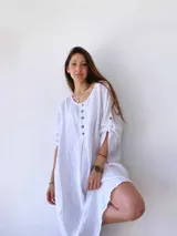 Vestido corto de lino en color blanco, con corte holgado, cuello redondo y cierre frontal parcial con botones. Presenta mangas tres cuartos con detalle de trabilla y botón.