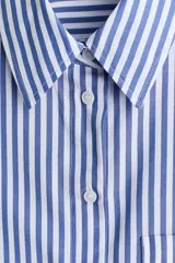 Camisa corta oversize de popelina de algodón con rayas verticales blancas y azules, cuello clásico, cierre frontal de botones, bolsillo en el pecho y mangas tres cuartos con puños anchos.