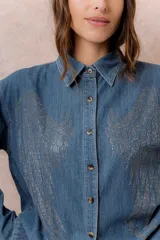 Camisa de denim liviano con calce relajado, cuello camisero clásico y cierre frontal con botones. Presenta una destacada aplicación de strass en el frente formando un diseño de alas.