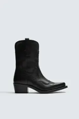 Bota estilo cowboy de caña media, confeccionada en piel negra con costuras decorativas en la caña y el empeine. Presenta una horma en punta, tacón ancho y tiradores laterales.