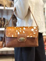 Cartera de cuero marrón con solapa de pelo vacuno en tonos marrón y blanco, cierre metálico de giro frontal y correa regulable.