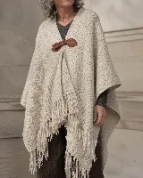 Ruana confeccionada en lana merino color beige, de diseño amplio y envolvente con flecos en el borde inferior y un broche artesanal de cuero en el frente.