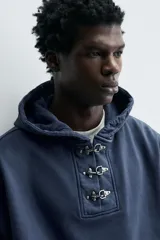 Sudadera hoodie relaxed fit color azul, confeccionada en tejido de algodón. Presenta capucha y cierre frontal con alamares metálicos, manga larga y acabados en rib.