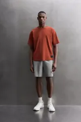 Camiseta de ajuste relajado (relaxed fit) color terracota, confeccionada en tejido técnico. Presenta cuello redondo, mangas ranglán cortas y un parche negro con logo en el bajo.