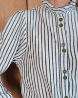 Camisa de manga larga con estampado de rayas verticales en tonos crudo y beige. Presenta cuello alto con volados, cierre frontal con botones dobles y puños con volados.