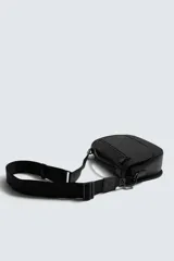 Bandolera de textura granulada color negro, con solapa frontal que incluye un cierre de cremallera. Cuenta con un asa superior fija y una correa de hombro ajustable y extraíble. El interior dispone de un bolsillo con cierre y un bolsillo plano.