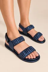 Sandalias de cuero vacuno metalizado azul marino, con suela plana, dos tiras frontales fruncidas y tira ajustable al tobillo con cierre de velcro.