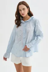 Camisa de algodón con detalles de broderie, volados y cuello a la base. Presenta diseño holgado, cierre frontal con botones y mangas largas con puños fruncidos.