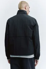 Campera rompevientos de nylon color negro, repelente al agua, con cuello elevado, cierre frontal y bolsillos laterales. Cuenta con forro de malla y puños con elástico.