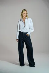 Camisa blanca de corte mullet, más larga en la parte posterior, con cuello camisero, bolsillo aplicado en el pecho, mangas largas con puños anchos y detalle de botones en la espalda.