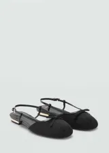 Zapato tipo ballerina destalonado con tira ajustable al tobillo, confeccionado en material textil negro con detalle de lazo decorativo en la punta redonda y tacón bajo con detalle metálico dorado.