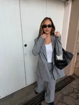 Conjunto de dos piezas compuesto por un blazer y un pantalón de corte sastrero, confeccionados en tela de algodón con estampado de rayas verticales blancas sobre fondo gris. El blazer presenta solapas clásicas y cierre frontal, mientras que el pantalón es de tiro medio y pierna recta.