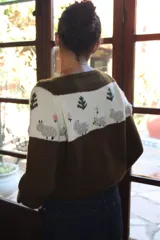Sweater tejido a mano en punto jersey, color marrón con franja superior en color natural y cuello redondo. Presenta bordados artesanales en relieve de ovejas, árboles y flores en el pecho.