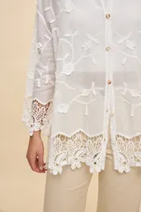 Camisa blanca de calce relajado, con cuello clásico, mangas 3/4 y cierre frontal de botones. Presenta bordados florales en relieve y detalles de crochet calado en los puños y el ruedo.