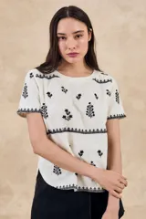 Remera de algodón color blanco con bordados florales y geométricos en color negro. Presenta cuello redondo, mangas cortas y un diseño con franjas horizontales bordadas.