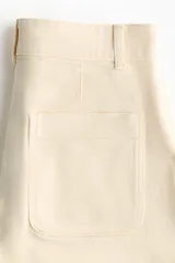 Short de tiro alto en denim de algodón levemente elástico, color beige, con bolsillos delanteros de parche con ribete y cierre frontal con botón.