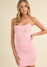 Vestido corto ajustado al cuerpo, de estilo bodycon, con breteles finos y escote redondo. Presenta un diseño en color rosado con aplicaciones de pedrería brillante en forma de estrellas o destellos distribuidas por toda la prenda.
